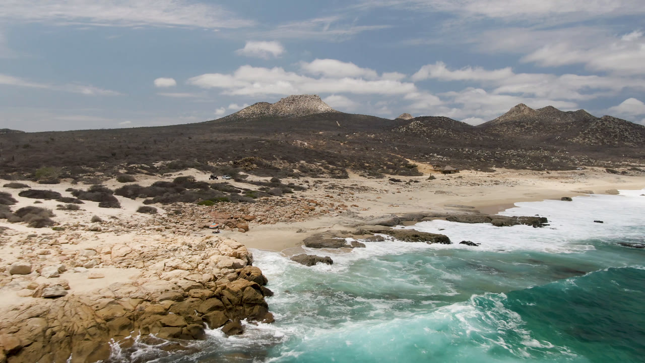 Rock outcrop beside bright surf with empty sand and hills in the background.