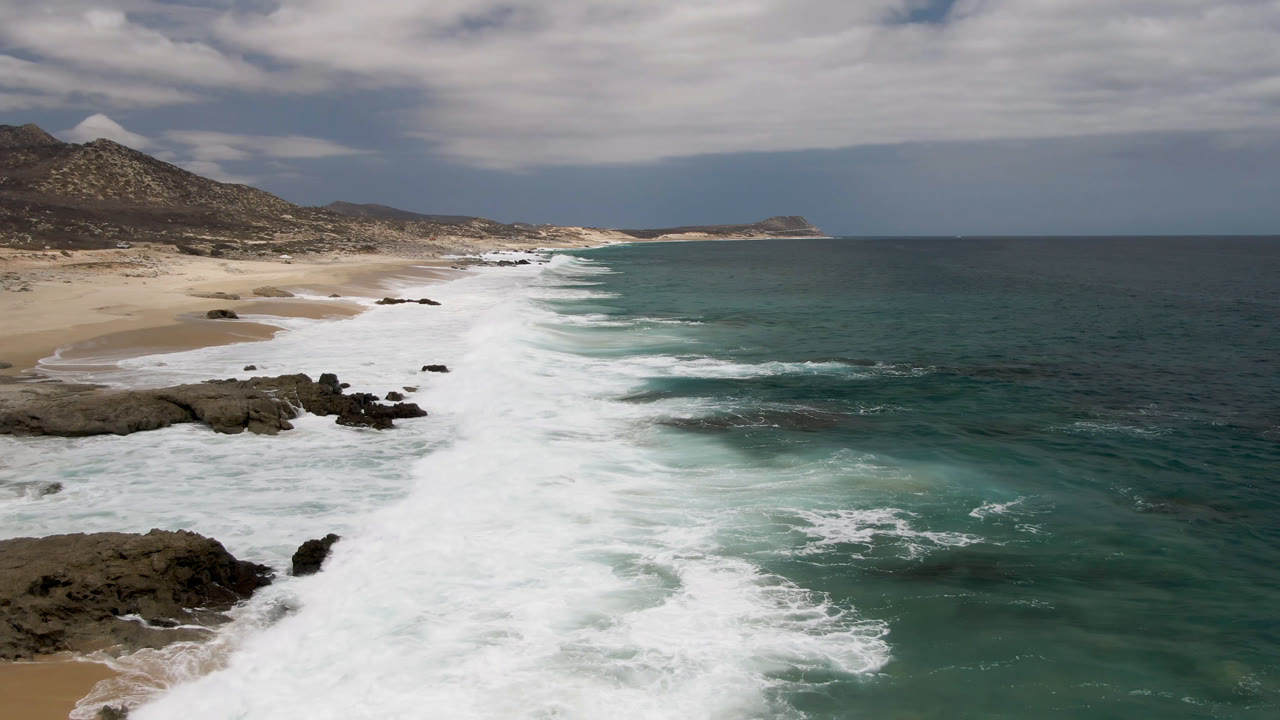 Laguna Beach in Baja California Sur, Mexico, seen from an elevated coastal angle as white surf spreads across teal water toward sand and dark rocks, with no visible people.