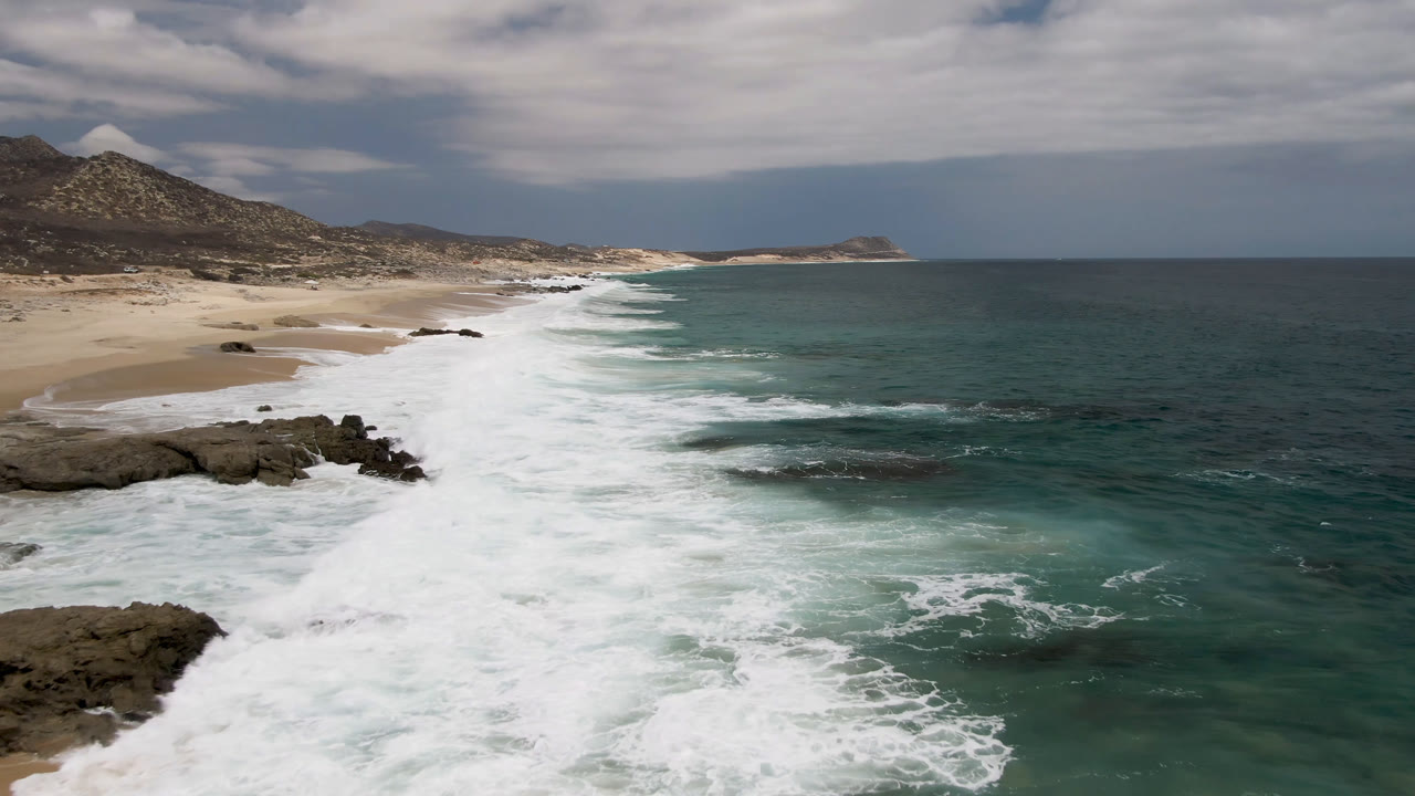 At 0:01, Laguna Beach surf forms a broad white diagonal along the sand while foreground rocks sit near foamy water and low hills rise behind the beach, with no visible people.