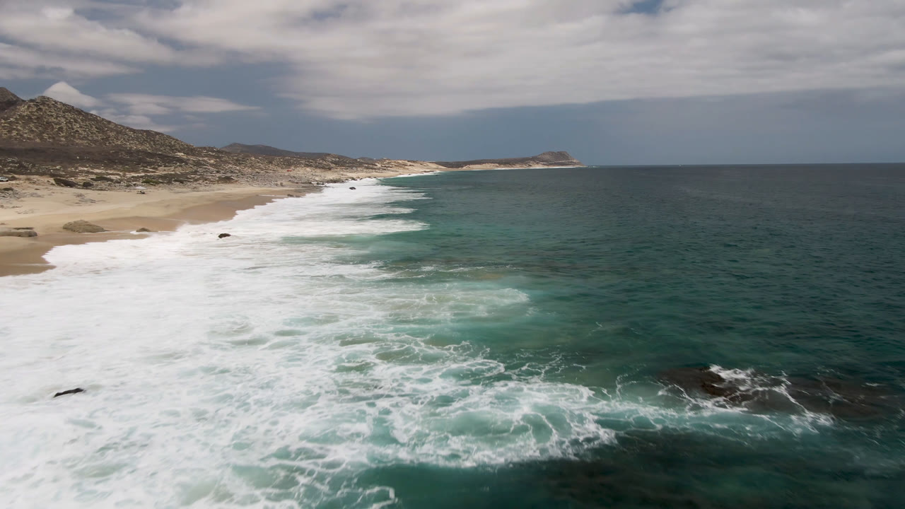 Elevated coastal view at Laguna Beach, Baja California Sur, with white surf spreading over sand, teal water to the right, low hills inland, and no visible people.