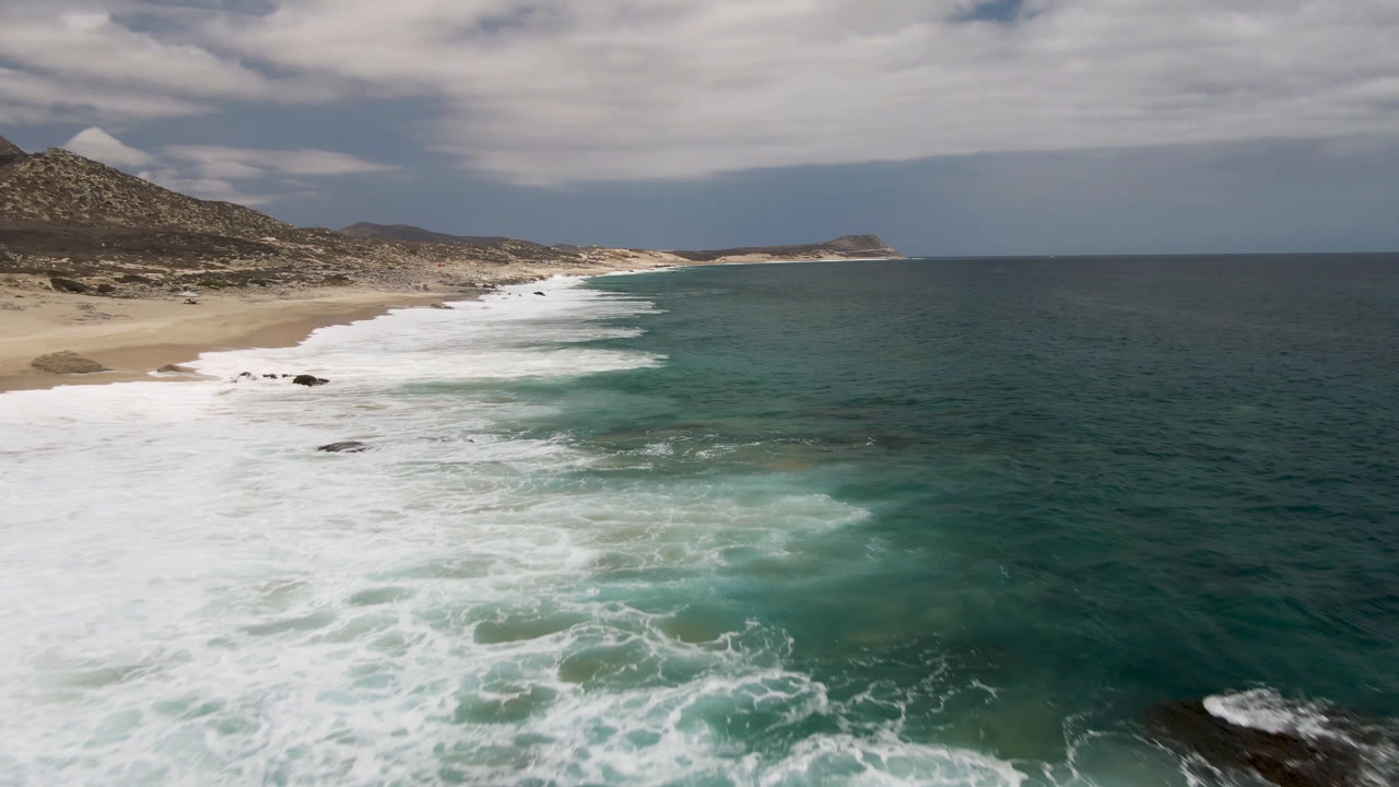 Laguna Beach, Baja California Sur shoreline with a broad band of white foam crossing the sand, darker rocks near the surf, open teal water, and no visible people.