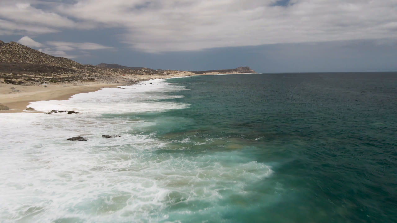 Wide aerial coastline with white surf, sandy beach, rocky patches, and cloudy sky.