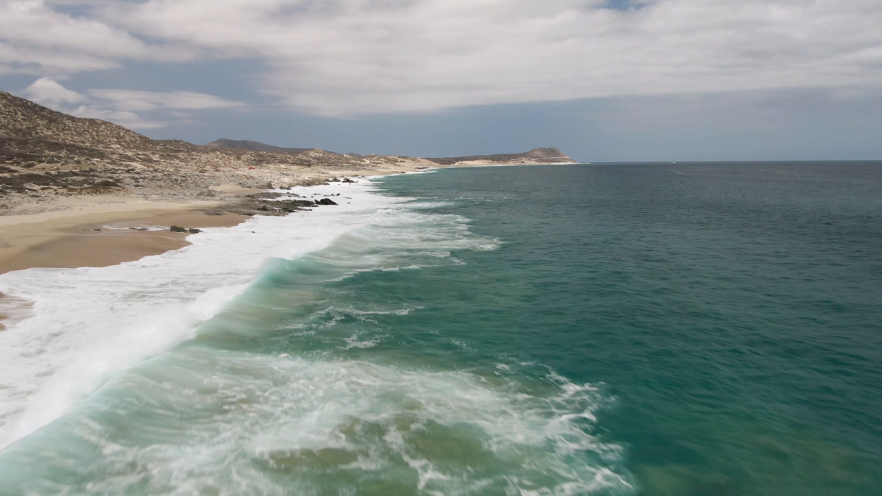 Aerial view of Laguna Beach in Baja California Sur, Mexico, with white surf crossing a sandy shoreline, rocky hills at left, open teal ocean at right, and no visible people.