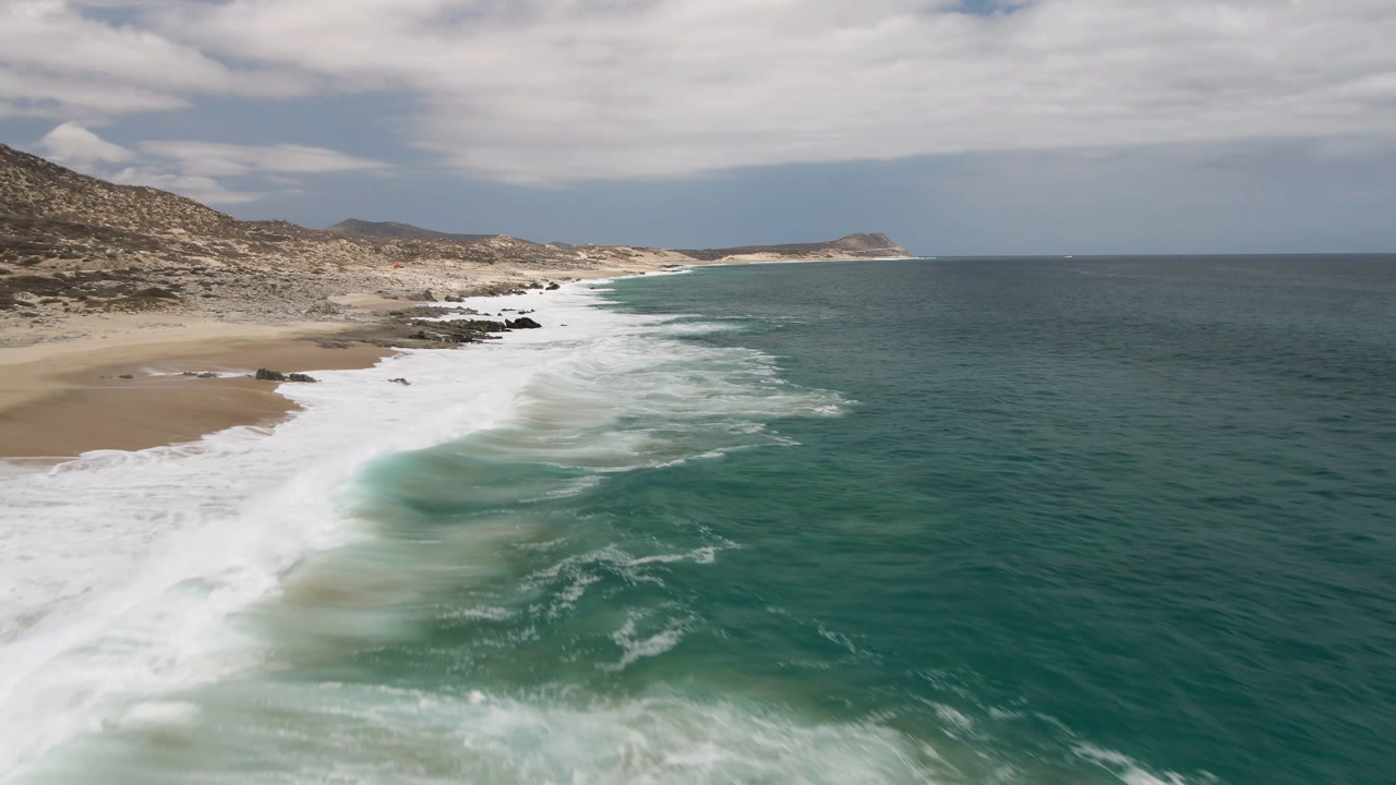 Laguna Beach surf rolls diagonally across the sand in Baja California Sur, Mexico, with foamy water nearer the center, dry coastal hills to the left, and no visible people.