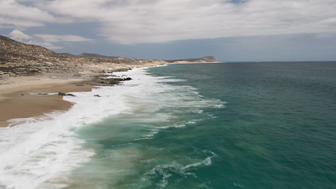 Broad ocean and foamy shoreline seen from above under a cloudy sky.