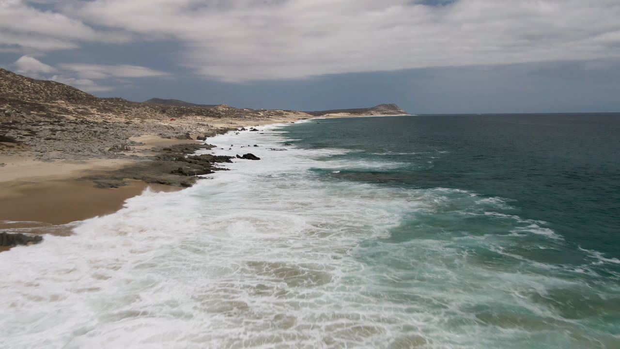 Aerial view of Laguna Beach in Baja California Sur, Mexico, with white surf spreading over rocks beside tan sand, teal ocean to the right, cloudy sky above, and no visible people.