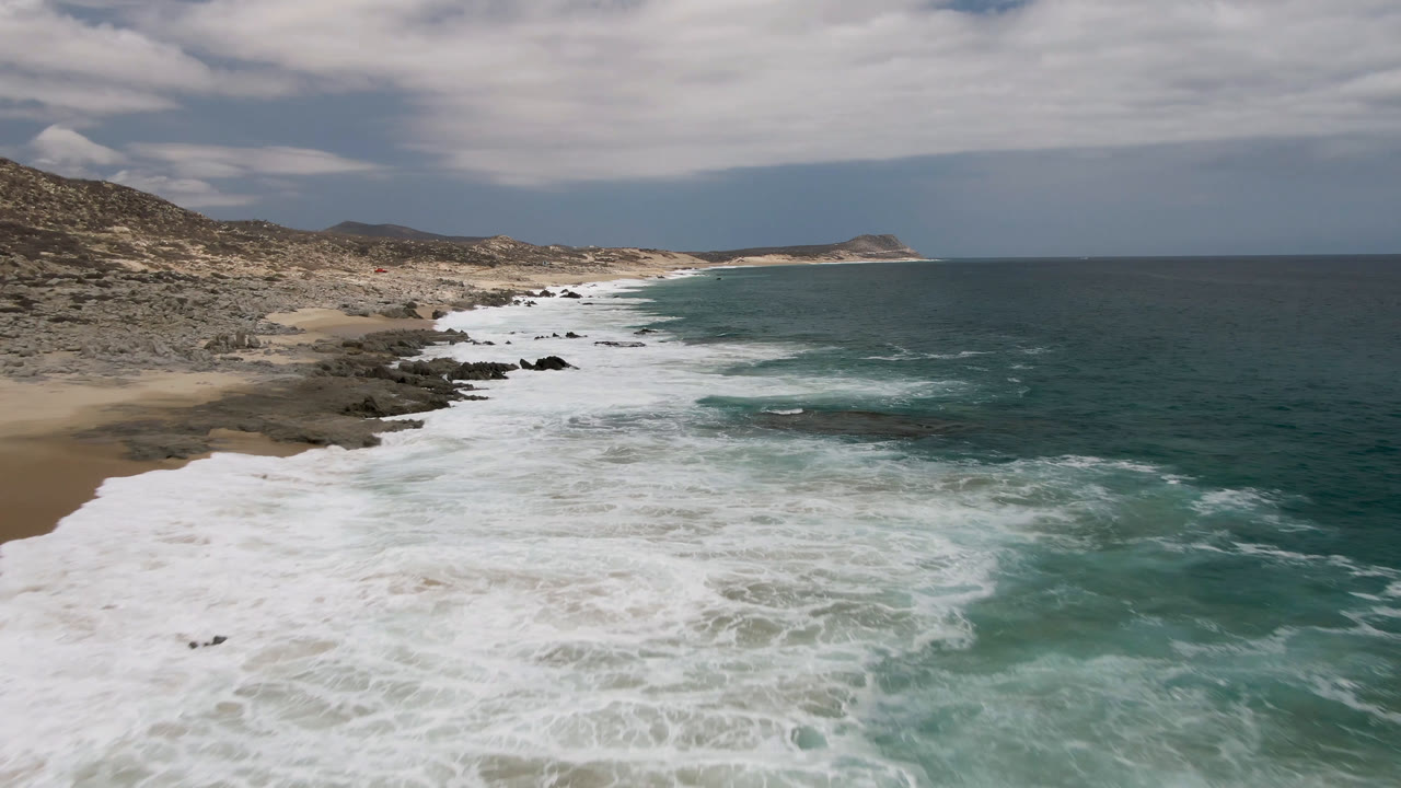 Laguna Beach shoreline from above, with foamy waves advancing over dark rocks, a small sandy cove on the left, low hills behind the coast, open ocean on the right, and no visible people.