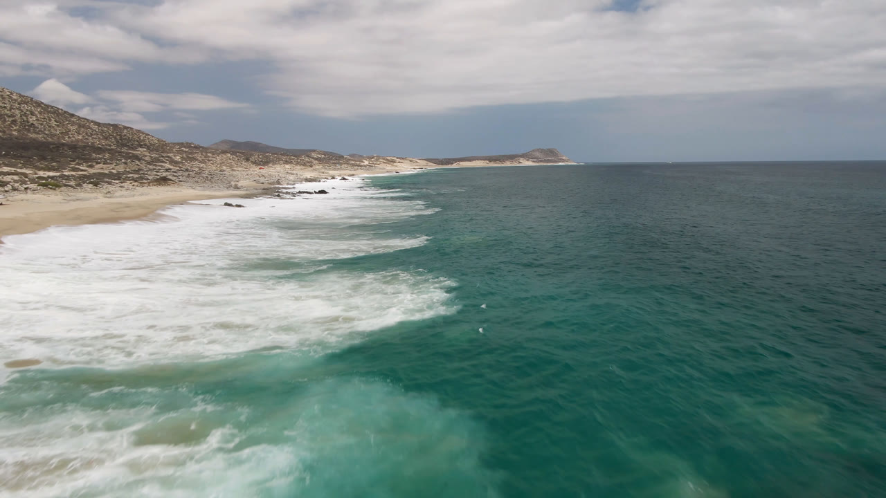 Aerial frame over Laguna Beach in Baja California Sur with white surf spreading across the left beach, turquoise water in the foreground, low hills inland, and no recognizable people visible.