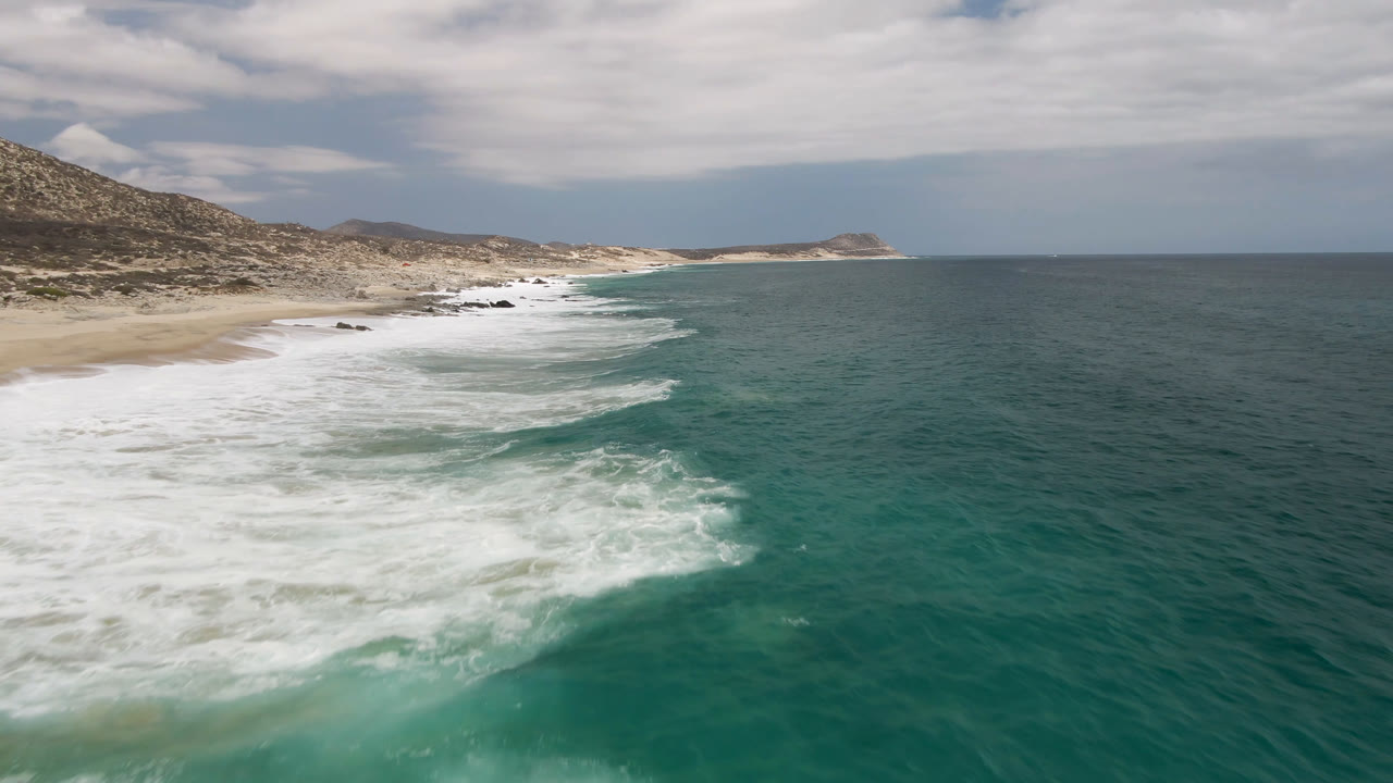 Wide aerial view of Laguna Beach in Baja California Sur as a long band of whitewater follows the sandy curve, with darker ocean to the right and low coastal hills under clouds.