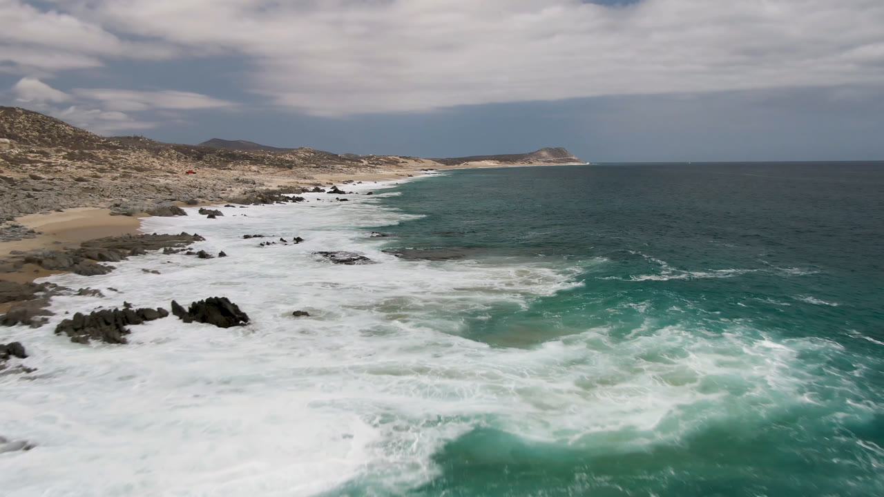 Elevated view of Laguna Beach in Baja California Sur, Mexico, with white surf spreading over dark rocks, turquoise water, sandy shore, low hills, cloudy sky, and no visible people.