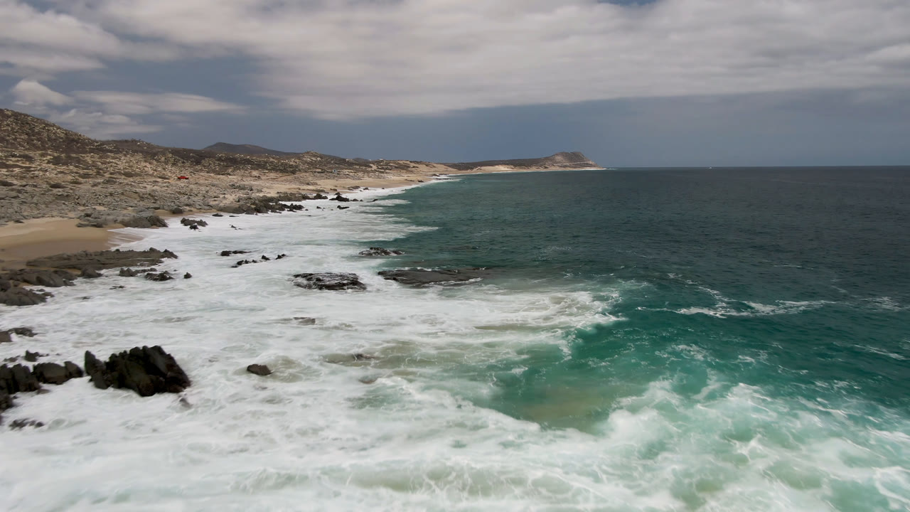 Laguna Beach coastline in Baja California Sur, Mexico, with foamy waves curling around nearshore rocks, a pale sandy beach to the left, open ocean to the right, and no visible people.