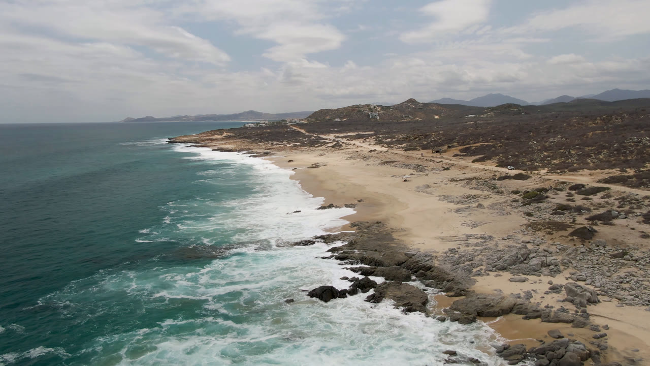 Aerial view over Laguna Beach in Baja California Sur, Mexico, with turquoise ocean, white surf, pale sand, rocky shoreline, scrub hills, and no visible people.