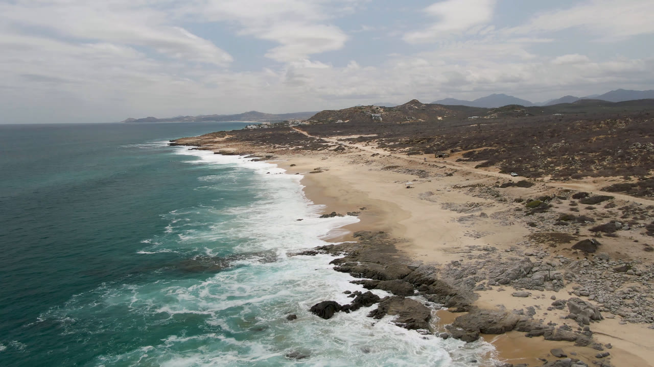 Drone-height view of Laguna Beach, Baja California Sur, where a band of whitewater curls onto dark rocks and tan sand beneath a bright clouded sky, with no visible people.