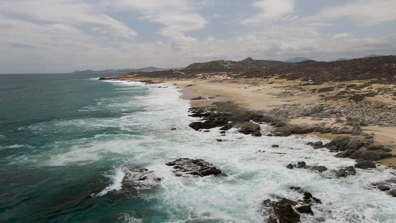 Aerial frame over Laguna Beach, Baja California Sur, with teal water, white surf, dark shoreline rocks, pale sand, scrubby hills, and no visible people.