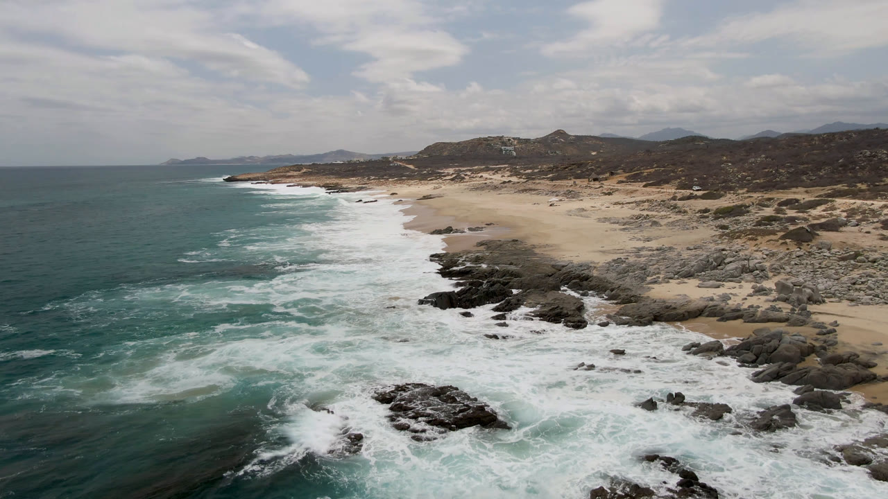 Laguna Beach, Baja California Sur, appears from an aerial angle as foam spreads around black rocks beside a wide sand beach, with no visible people.