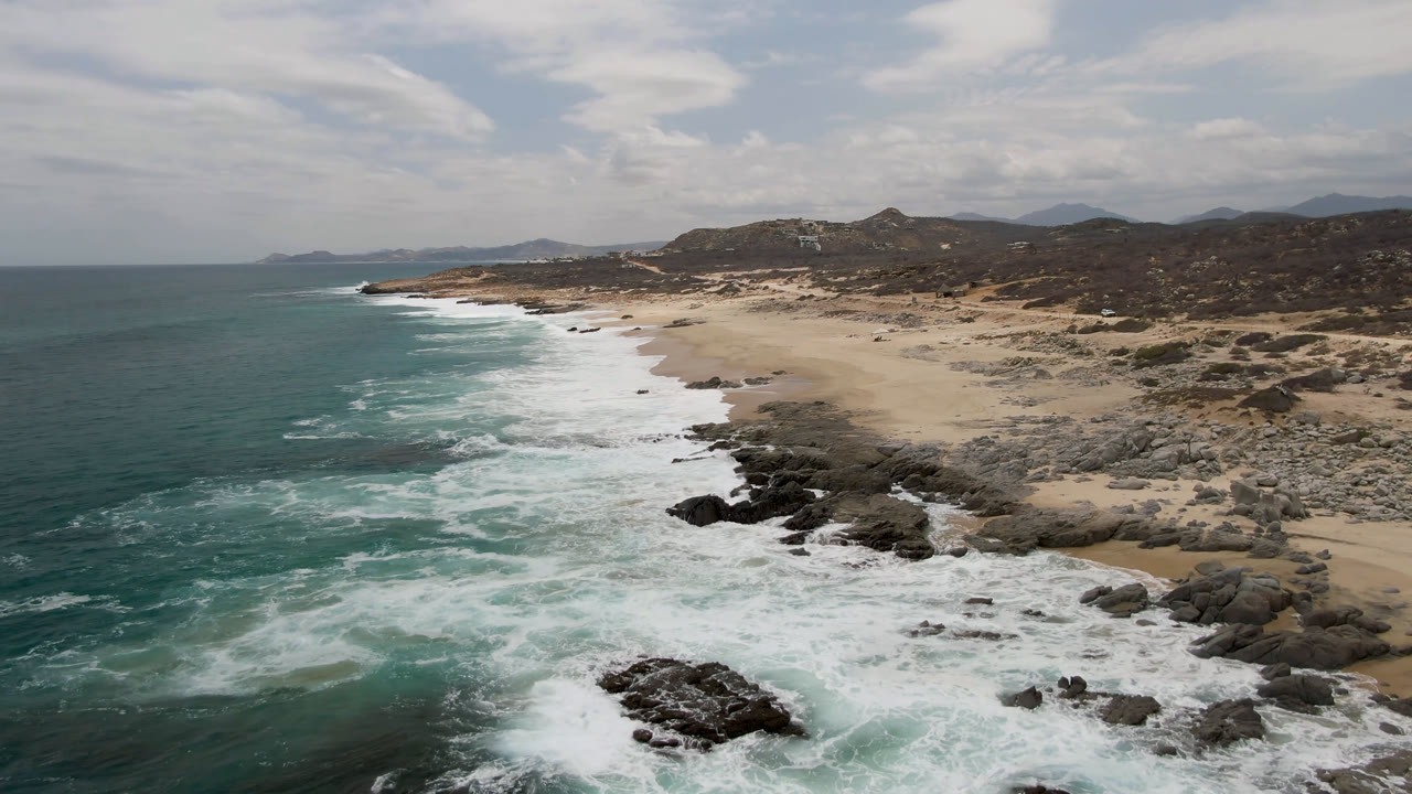 A wider aerial view of Laguna Beach, Baja California Sur, shows the coastline receding into the distance with surf bands, rocky outcrops, sand, hills, and no visible people.