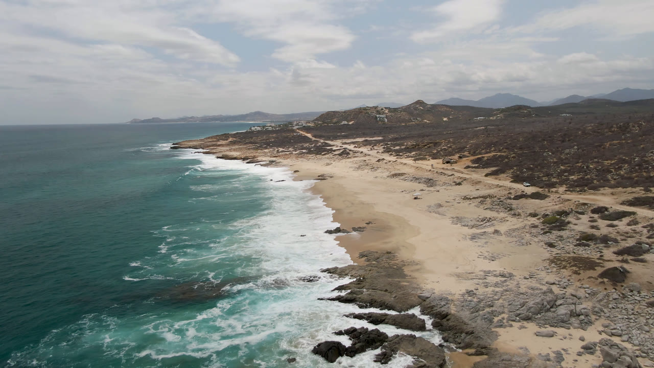 Opening aerial view of Laguna Beach, Baja California Sur, with turquoise ocean on the left, white surf meeting a sandy and rocky shore, dry hills inland, and no visible people.