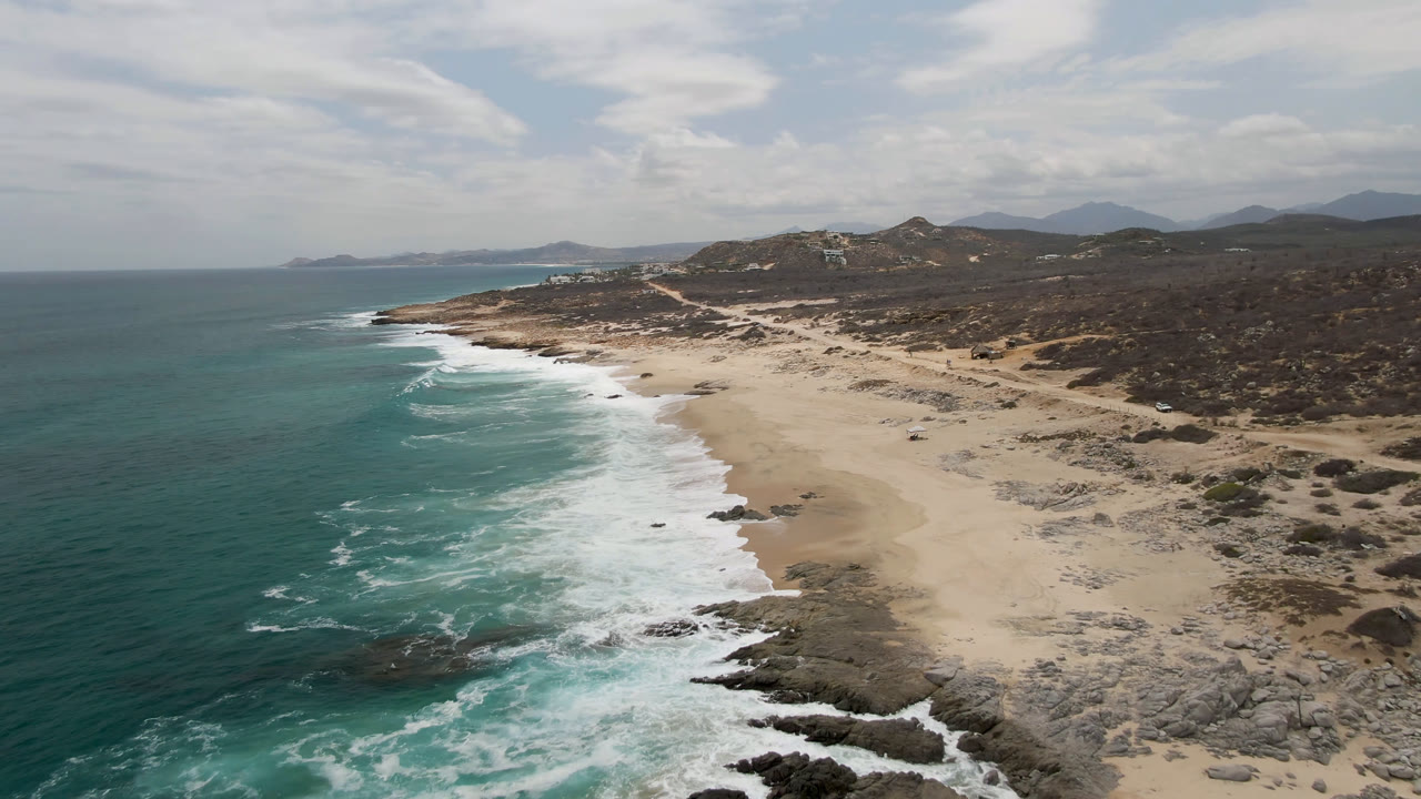 Second-frame aerial view of Laguna Beach, Baja California Sur, showing longer surf bands along the left edge, pale sand through the center, rocky shoreline below, inland hills, and no visible people.