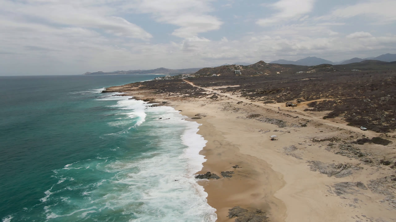 Aerial view of Laguna Beach in Baja California Sur with turquoise water on the left, white surf crossing the sand, scrubland inland, and no visible people.