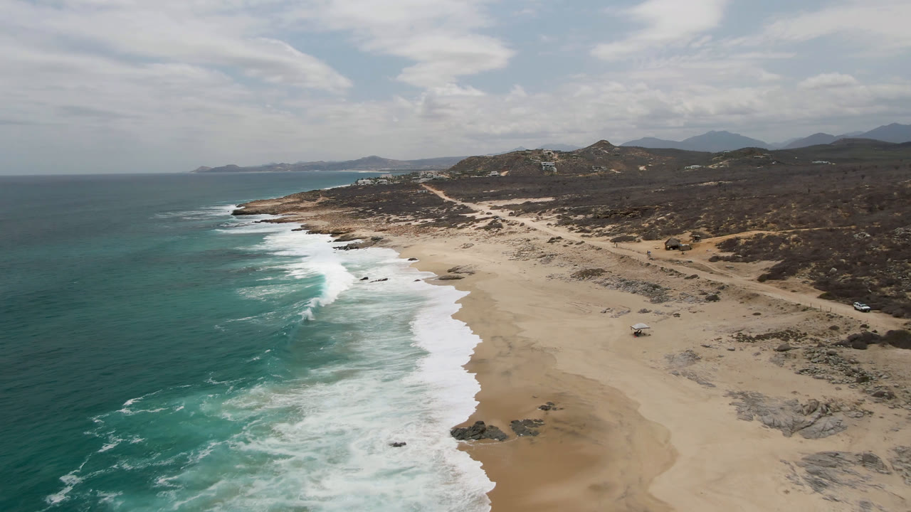 Aerial frame over Laguna Beach, Baja California Sur with a longer strip of foamy surf, pale sand in the center, inland scrub and hills beyond, and no visible people.
