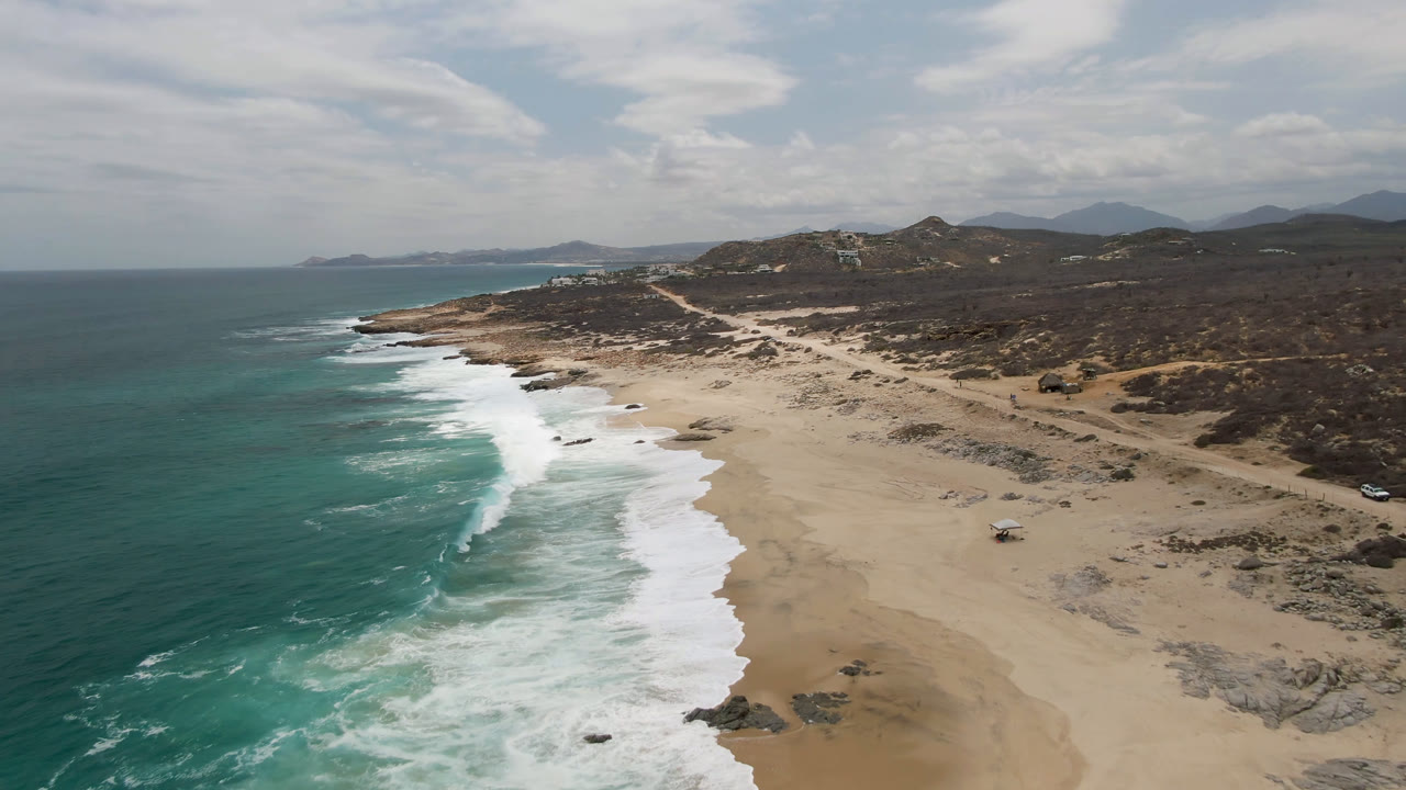 Drone view of curved surf, sandy shoreline, scrubland, and distant hills.