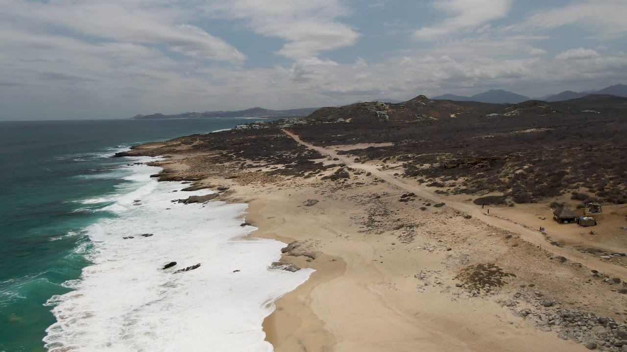 Aerial view over Laguna Beach in Baja California Sur, Mexico, with turquoise water at left, broad sand below, dark scrub inland, low hills on the horizon, and no visible people.