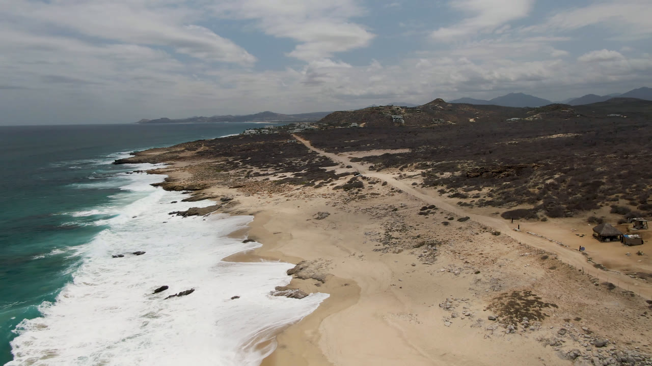 Drone-height view of Laguna Beach in Baja California Sur, Mexico, showing white surf curling farther up the sand, rock patches near the waterline, scrubland beyond the beach, and no visible people.