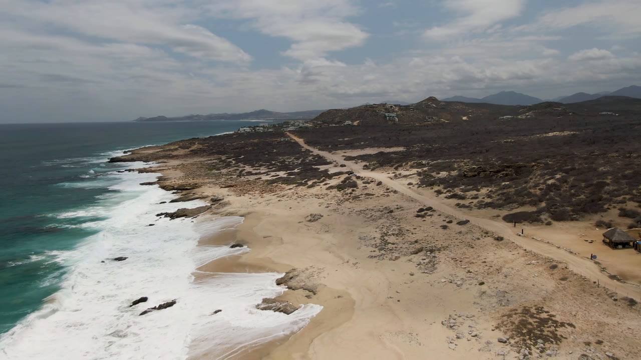 Aerial coastline view with foamy surf, sandy beach, a dirt road, scrubland, and distant hills.