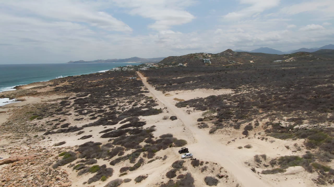 Aerial frame from Laguna Beach, Baja California Sur with a dark vehicle on the sandy road, scrub patches across the foreground, ocean surf to the left, hills beyond, and no visible people.
