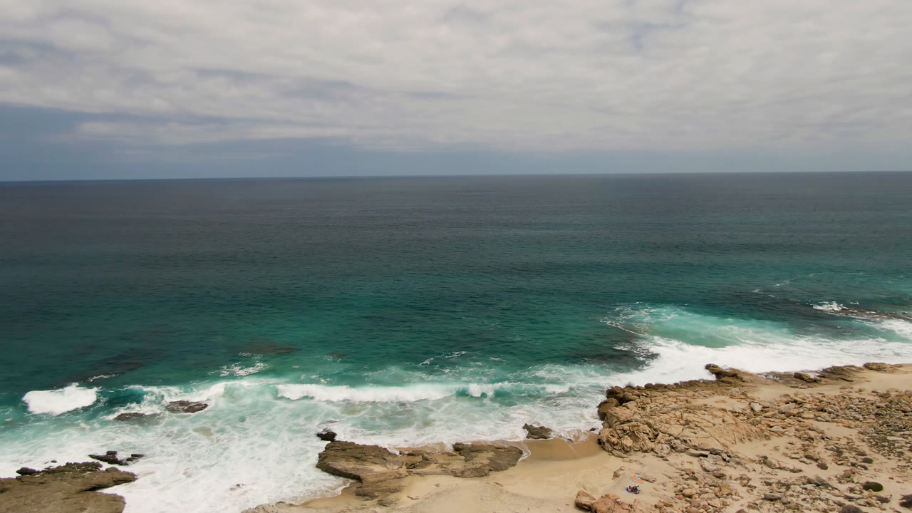 Wide elevated view of Laguna Beach in Baja California Sur with tan rocks, sandy shore, teal ocean, white surf, cloudy sky, and a tiny distant person near the lower shoreline.