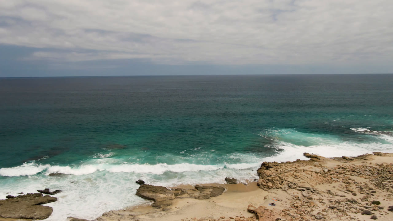 Laguna Beach shoreline from above, showing waves spreading across flat rock shelves, pale sand, turquoise water, a cloudy horizon, and a tiny distant person near the beach area.