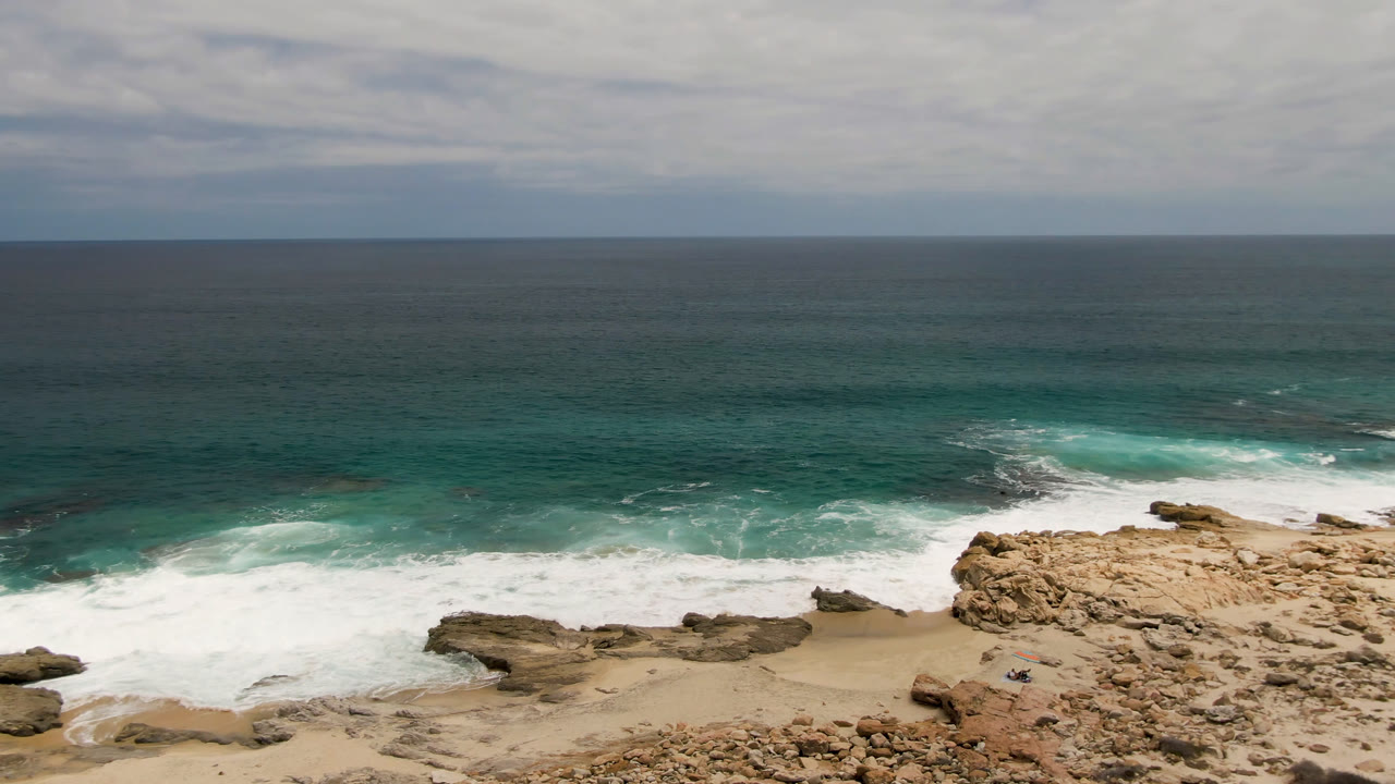 Wide elevated view of Laguna Beach, Baja California Sur, with tan rocks, a sandy pocket beach, white surf, turquoise water, and a tiny visible figure near the lower rocks.