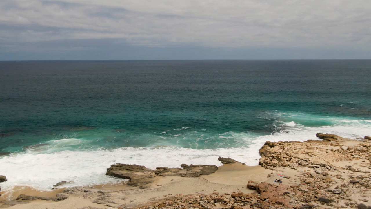 Laguna Beach, Baja California Sur shoreline with whitewater spreading across the left side of the beach, tan cliff rocks in the foreground, and a small figure or object near the lower rocks.