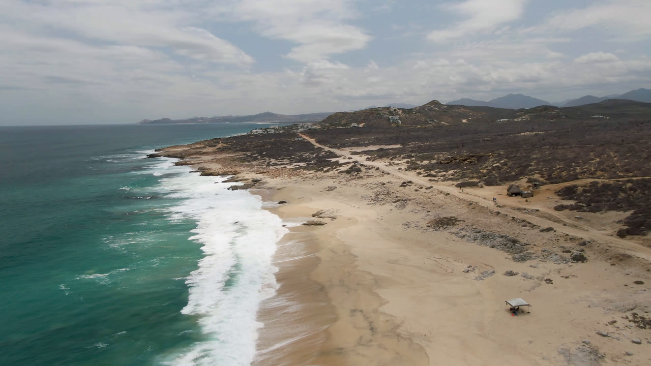Aerial view over Laguna Beach, Baja California Sur, with turquoise water, white surf, pale sand, inland scrub, distant hills, and no visible people at the start of the clip.