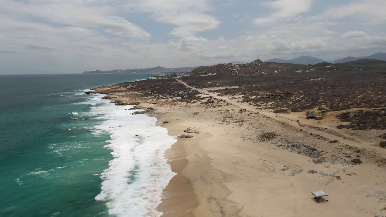 Drone view of Laguna Beach, Baja California Sur, with a thicker band of whitewater along the shore, open sand, scrubby inland terrain, distant hills, and no visible people.