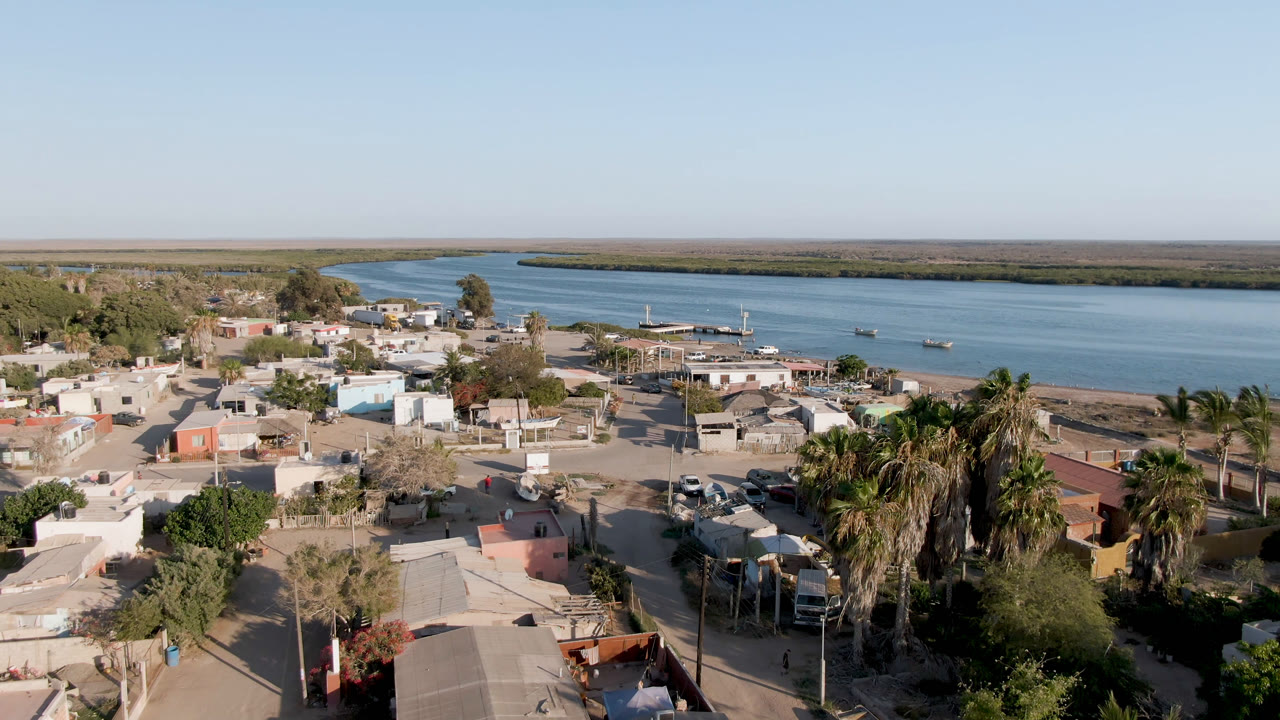 Opening aerial frame of Aldofo Lopez Mateos, Baja California Sur, with low buildings, sandy streets, palms, parked vehicles, a broad river, flat shoreline, and no visible people.