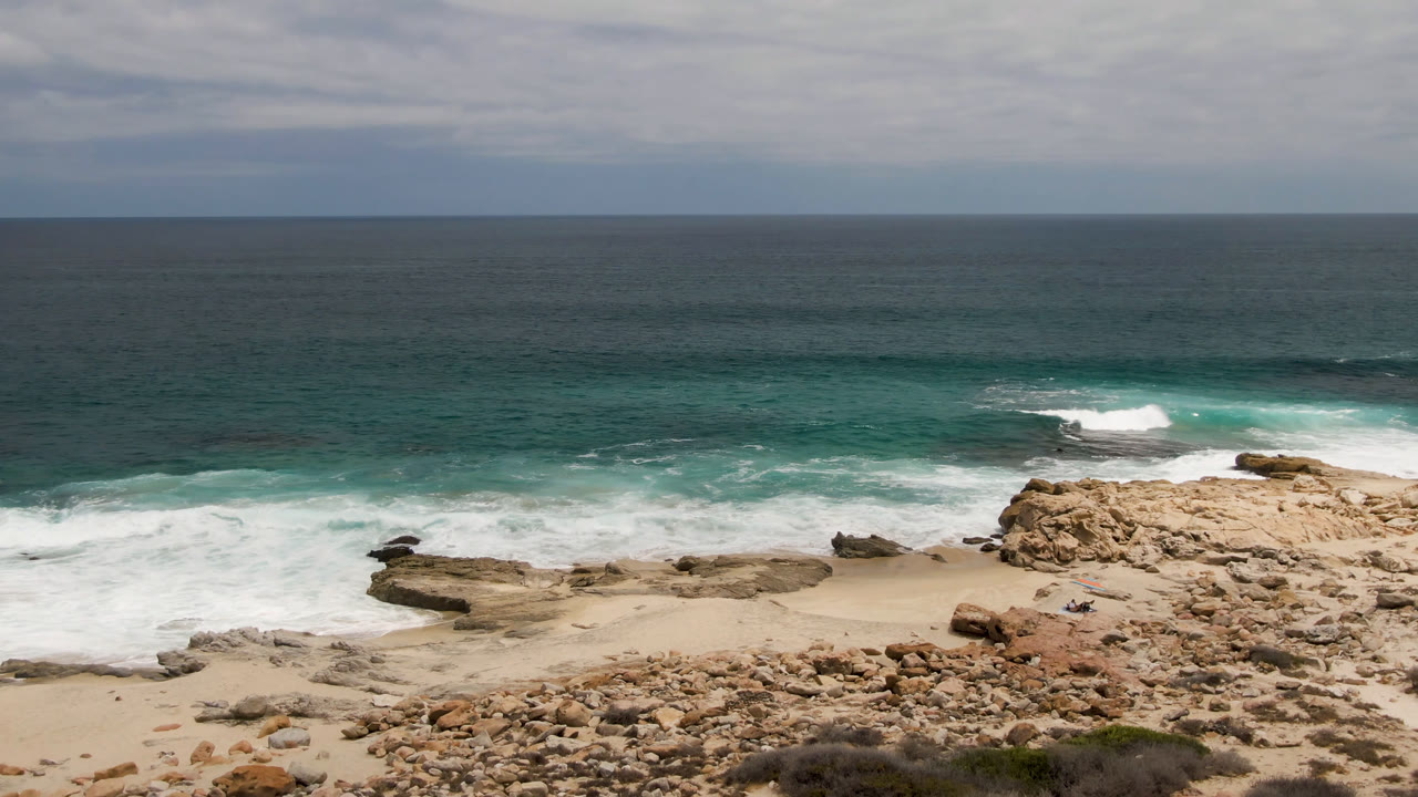 Wide elevated view of Laguna Beach in Baja California Sur with rocky sand, white surf, turquoise water, scrub in the foreground, and tiny distant beachgoers.