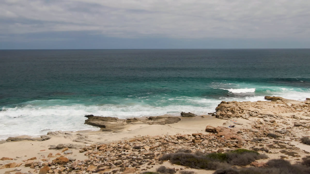 Laguna Beach shoreline in Baja California Sur with a central rock shelf, pale sand, foamy waves, dark teal water, and small distant figures near the beach.