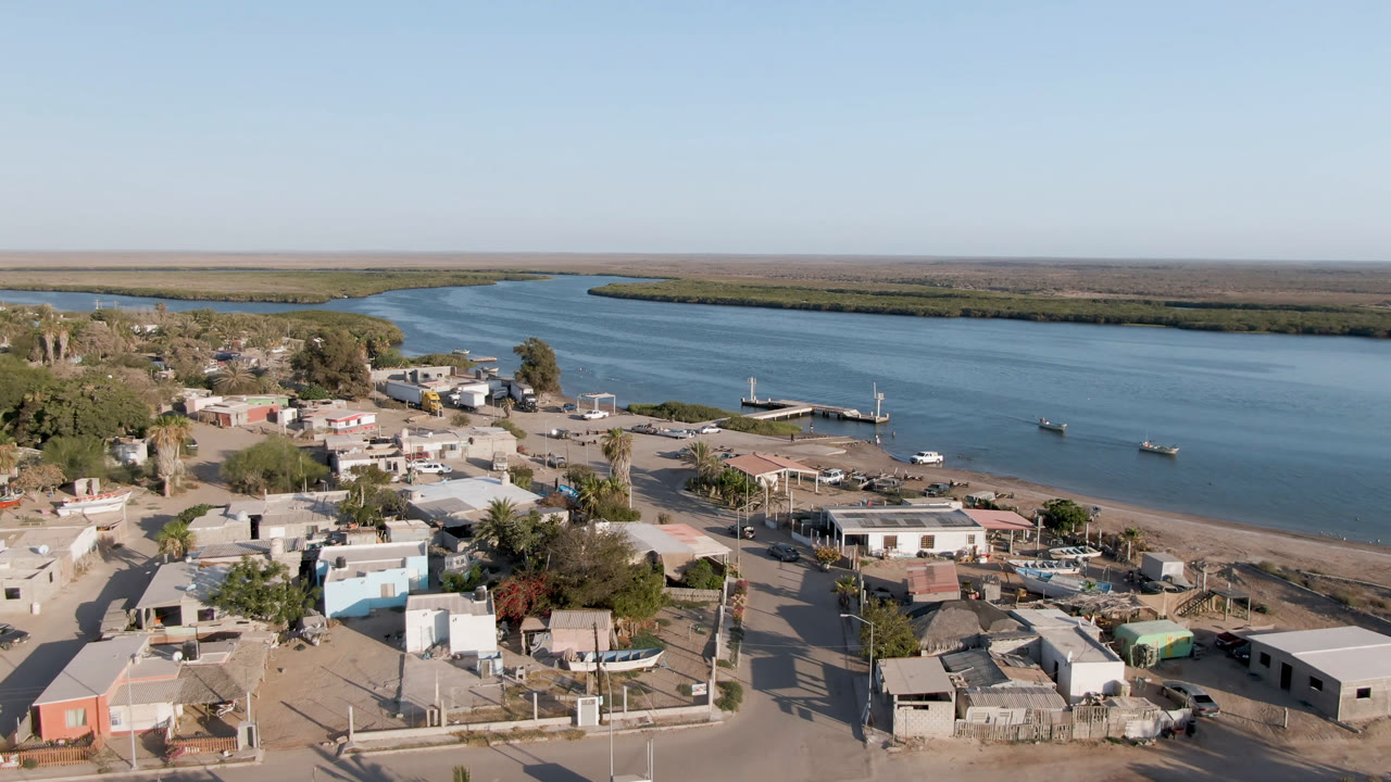 At 0:01 the aerial view in Aldofo Lopez Mateos looks across clustered rooftops and trees toward a dock and boats on the river, with no visible people.