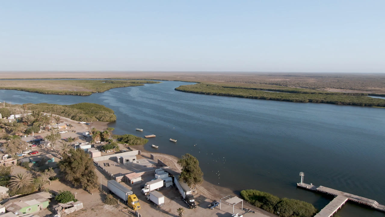 Aerial frame over Aldofo Lopez Mateos, Baja California Sur, with a dock in the lower right, low buildings and trees at the shoreline, open blue water, and no visible people.