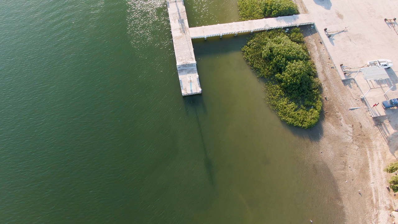 Aerial frame from Aldofo Lopez Mateos, Baja California Sur, Mexico, showing a concrete dock over green water beside a sandy shoreline with shrubs and no visible people.
