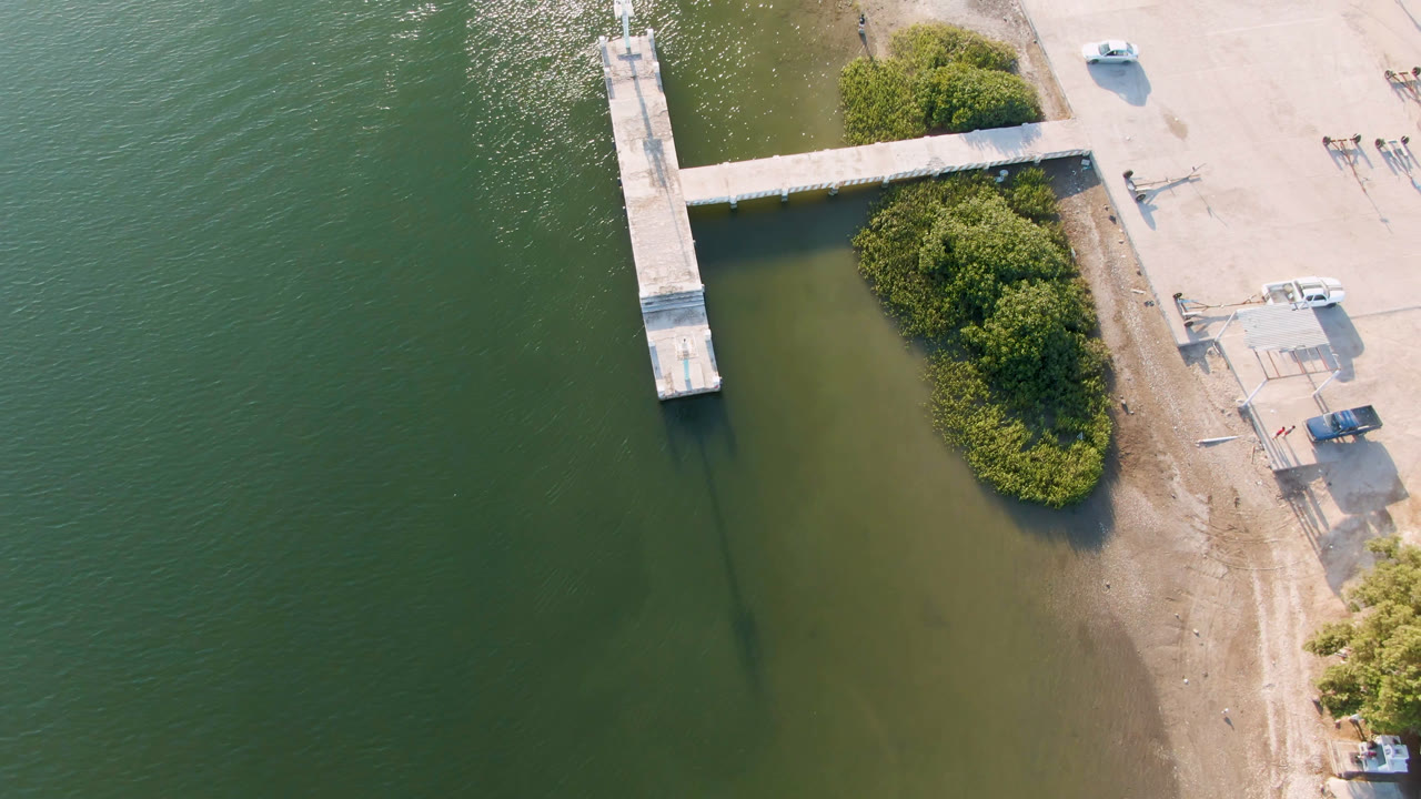 Aerial dock scene with green water, a sandy bank, shrubs, and vehicles on shore.