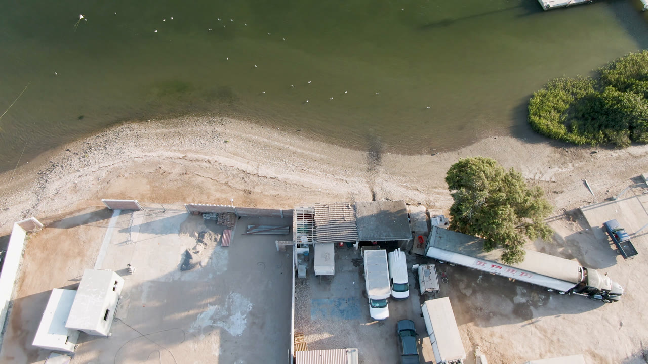 High aerial frame over Aldofo Lopez Mateos, Baja California Sur, showing green shallow water, a pale sandy shoreline, trees, parked trucks and trailers, and no visible people.