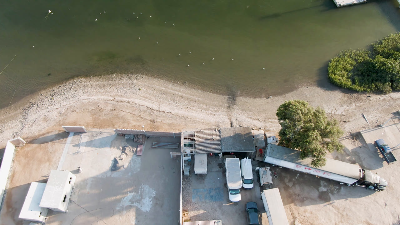 Aerial view in Aldofo Lopez Mateos, Baja California Sur with the curved sand edge below green water, a shrub island near shore, parked trucks, a trailer, and no visible people.