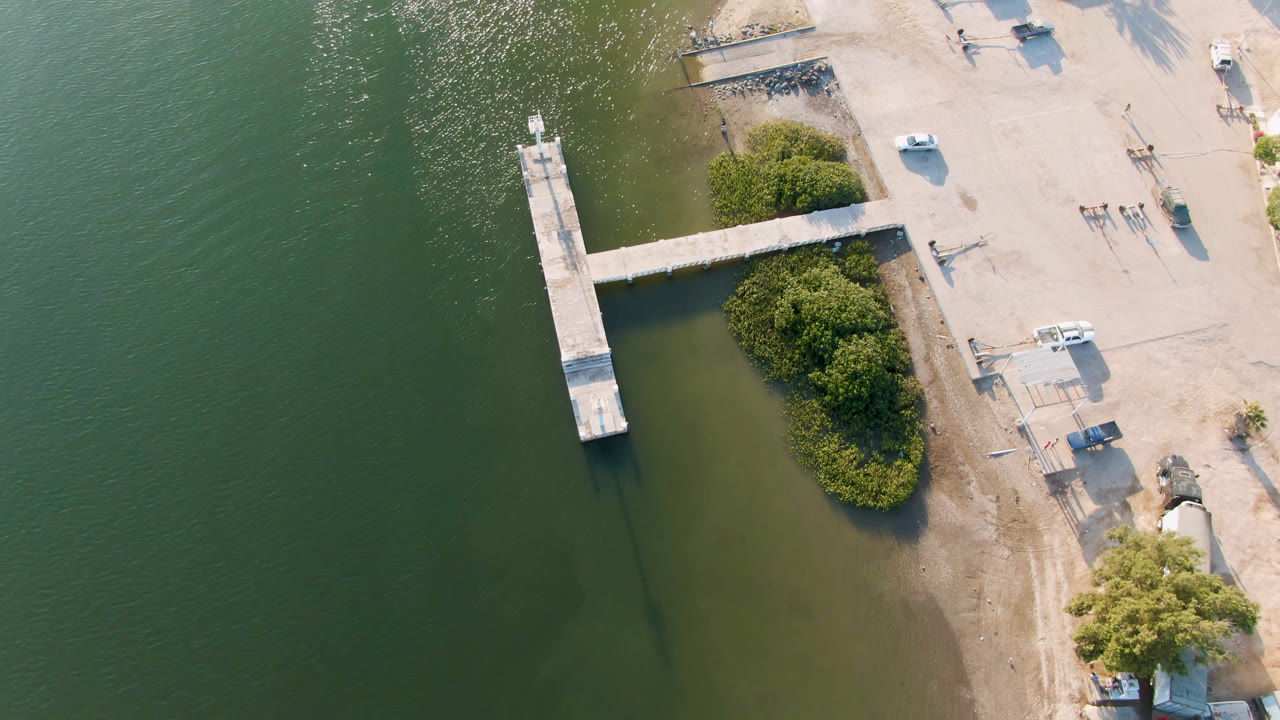 Overhead aerial frame in Aldofo Lopez Mateos, Baja California Sur, Mexico, showing green water, a narrow dock, shoreline vegetation, parked vehicles, small waterfront structures, and no visible people.