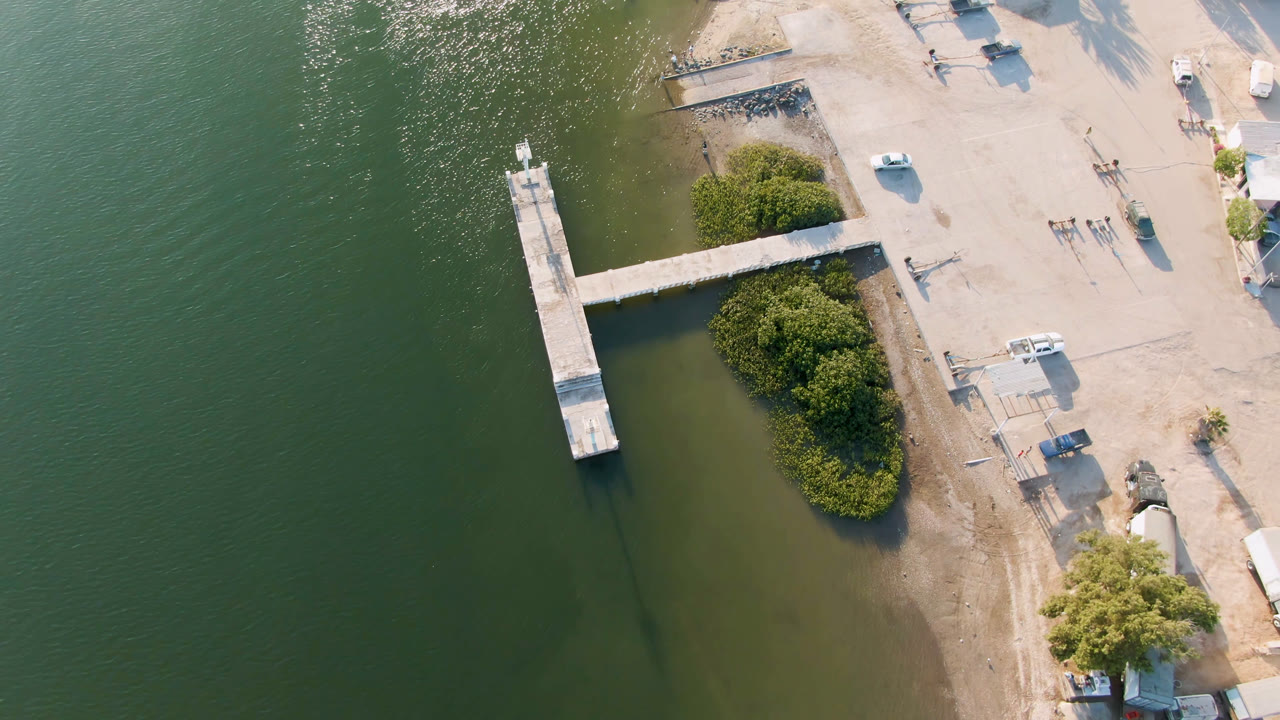 Aerial view over Aldofo Lopez Mateos with the dock centered between green water and a pale waterfront lot, with shrubs near the shore, parked vehicles, scattered structures, and no visible people.