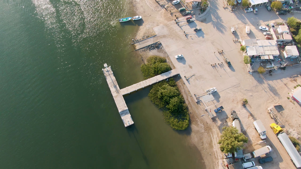Aerial view of green water, a wooden pier, sandy shore, buildings, trees, and parked vehicles.