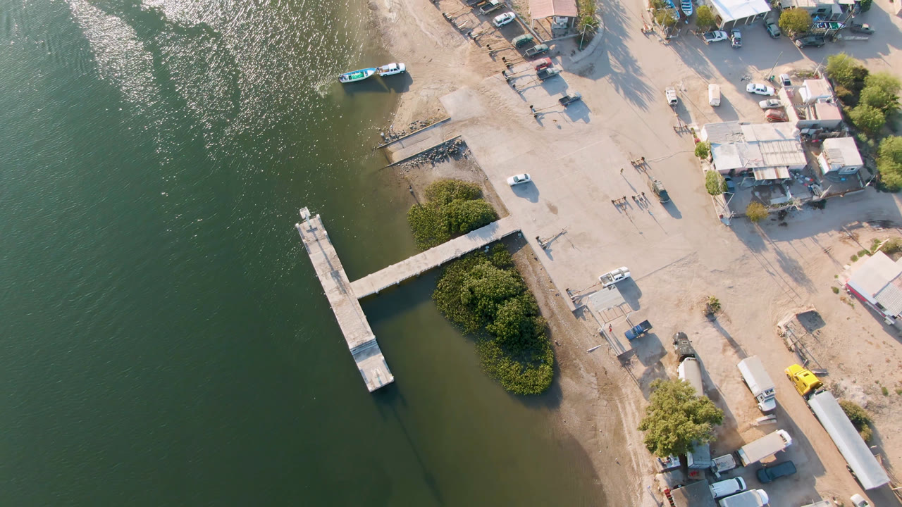 Second frame over Aldofo Lopez Mateos, Baja California Sur, with the pier angled across green water, a small boat near the upper shore, scattered buildings, parked vehicles, trees, and no visible people.