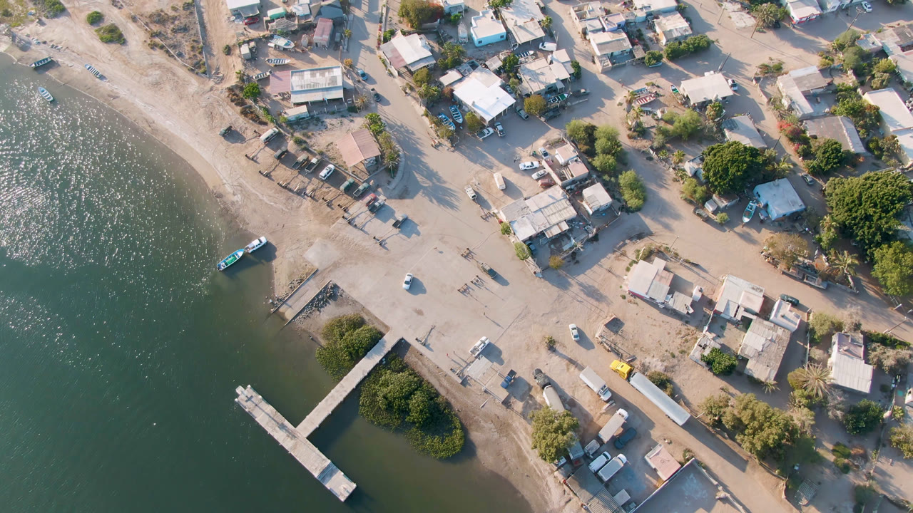 Second aerial frame over Aldofo Lopez Mateos, Baja California Sur, with the dock near the lower left, a small boat close to shore, rippled green water, scattered buildings, sandy roads, parked vehicles, trees, and no visible people.