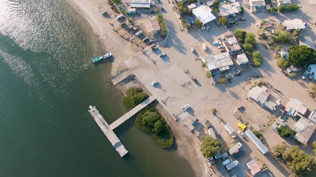 Aerial frame over Aldofo Lopez Mateos with the dock projecting into green water, a turquoise boat near the shoreline, dirt roads, parked vehicles, trees, low buildings, and no visible people.